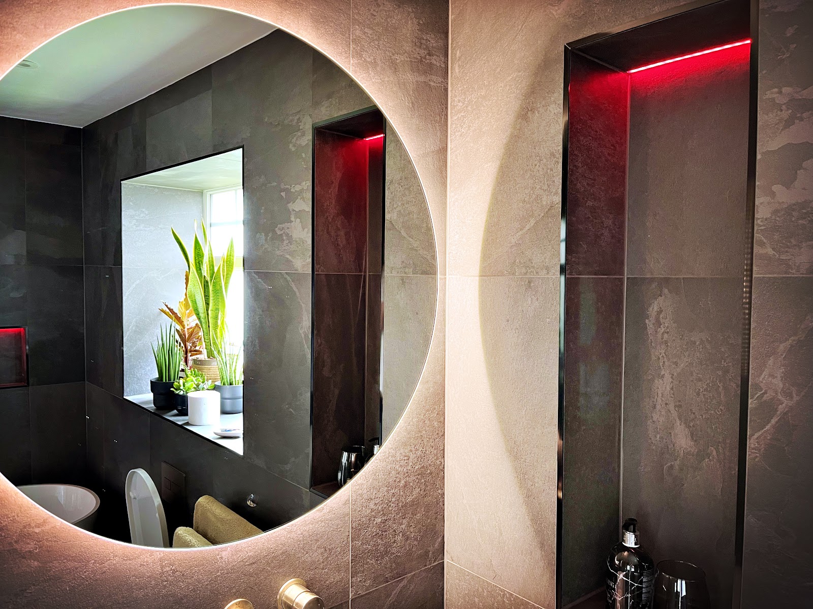 Round backlit mirror in a dark stone bathroom with red LED strip
